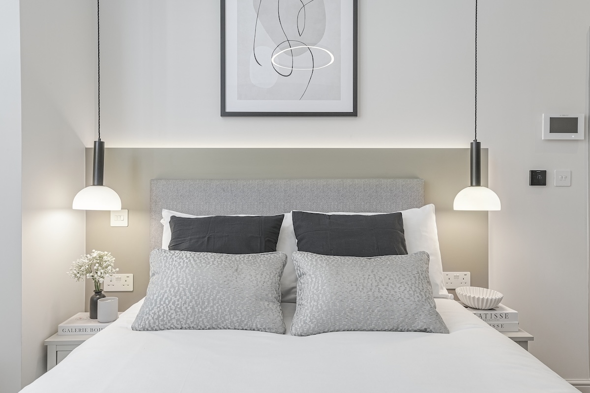 A modern bedroom with a neatly made bed, grey and white pillows, a grey headboard, hanging pendant lights on each side, books on bedside tables, and abstract wall art above the bed. The space is bright and minimalistic.