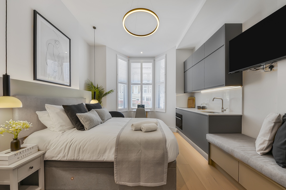 Modern studio bedroom with a large bed, grey headboard, bedside tables, wall art, plants, kitchenette, built-in bench, TV, and large bay window with white shutters. Warm, neutral tones and soft lighting create a cosy atmosphere.