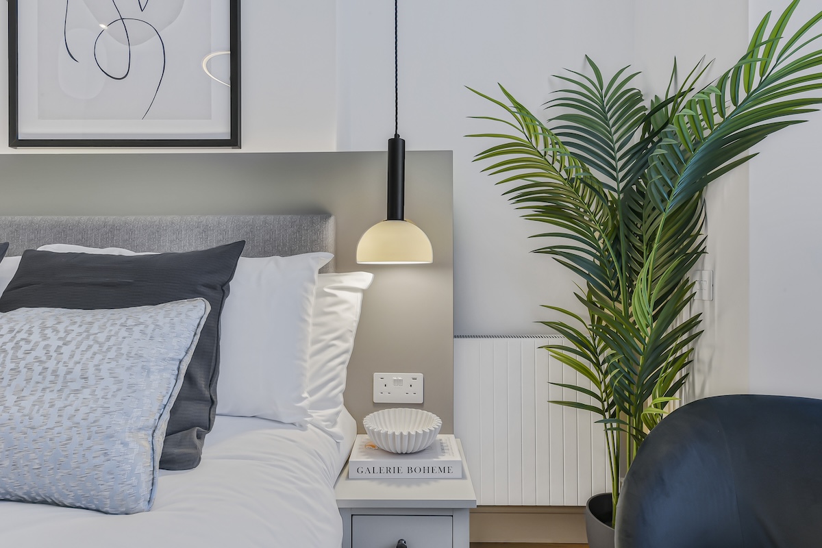 A modern bedroom corner with a neatly made bed, grey and white pillows, a small white bedside table holding books and a decorative bowl, a hanging lamp, and a tall green potted plant beside the bed.