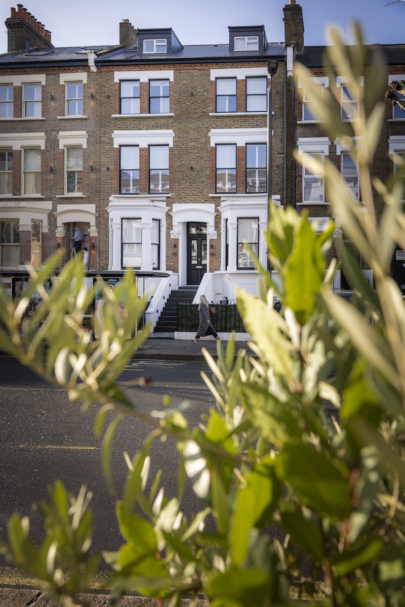 A person walks past a three-storey brick townhouse with white trim and black doors, viewed through leafy green branches in the foreground on a sunny day—introducing OneStay Maida Vale.