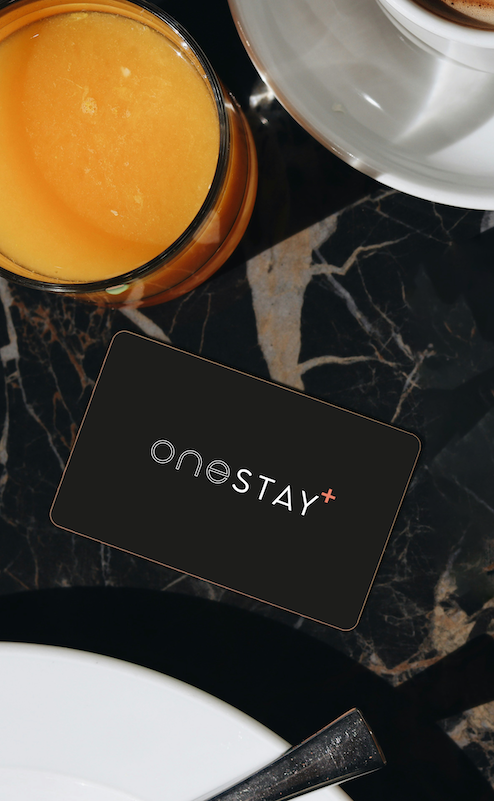 A black card with the text ONESTAY+ lies on a dark marble table, next to a glass of orange juice and a white plate with the edge of a cup visible.