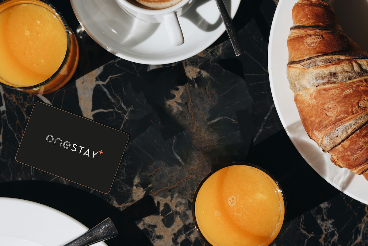 A dark marble table set with two glasses of orange juice, a white cup of coffee, a croissant on a plate, and a black OneStay+ card.