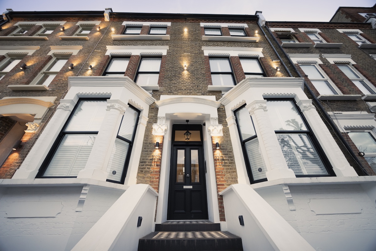 A wide-angle view looking up at the entrance of a renovated townhouse with brick and white trim, black door, white-framed windows, and outdoor wall lights. Steps lead up to the doorway.